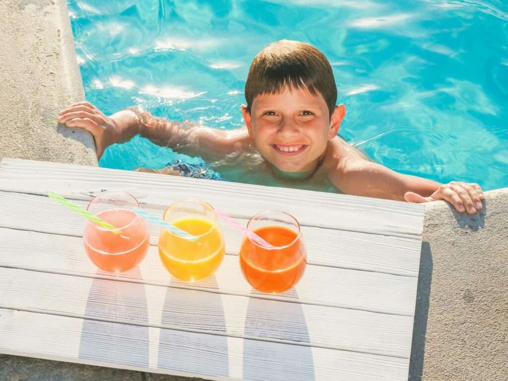 pexels-photo-8215125-8215125 Smiling child in a swimming pool with three colorful drinks on a sunny day.